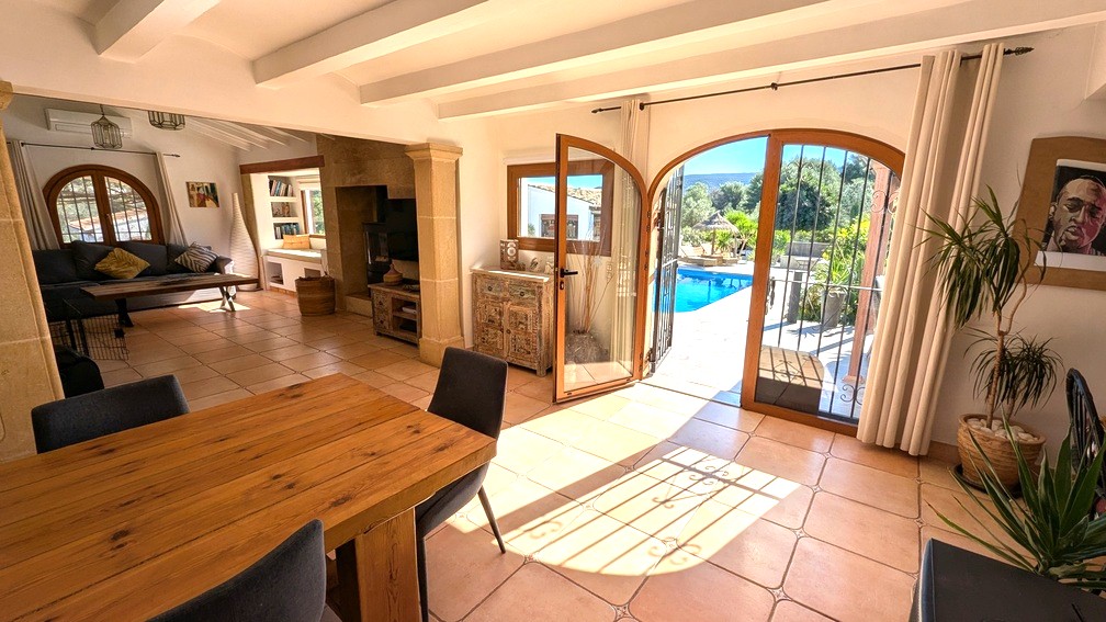 Salón-comedor de planta abierta con vigas blancas, pilares de piedra Tosca y puertas de arco que conectan el interior con la terraza y la piscina.