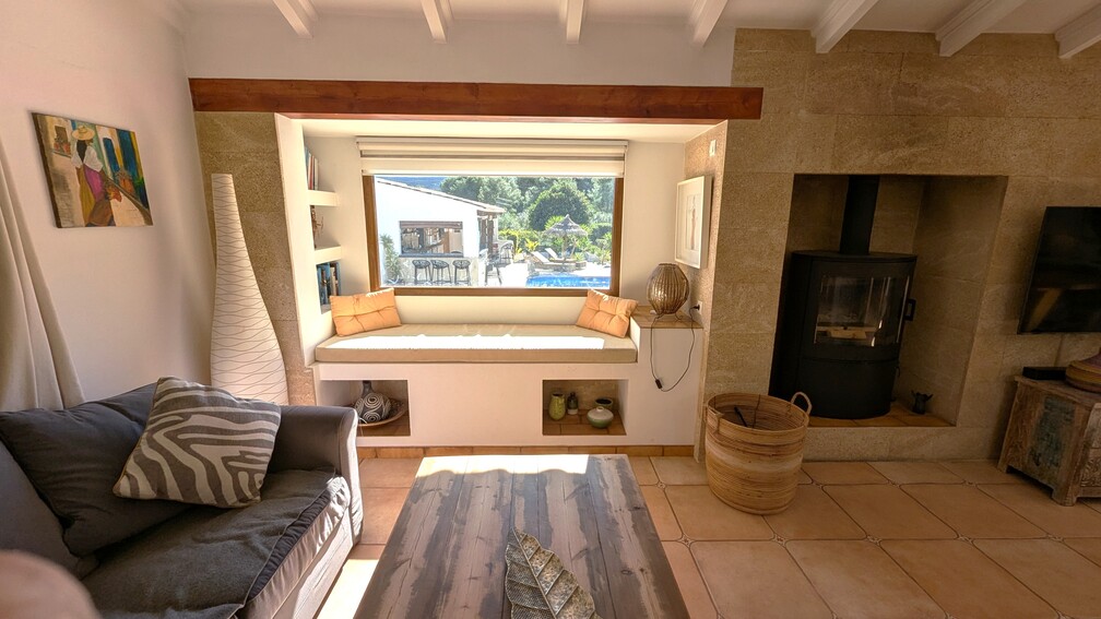 Salón con banco integrado bajo el ventanal con vistas a la piscina, chimenea de leña encastrada en pared de piedra tosca y techos con vigas vistas.