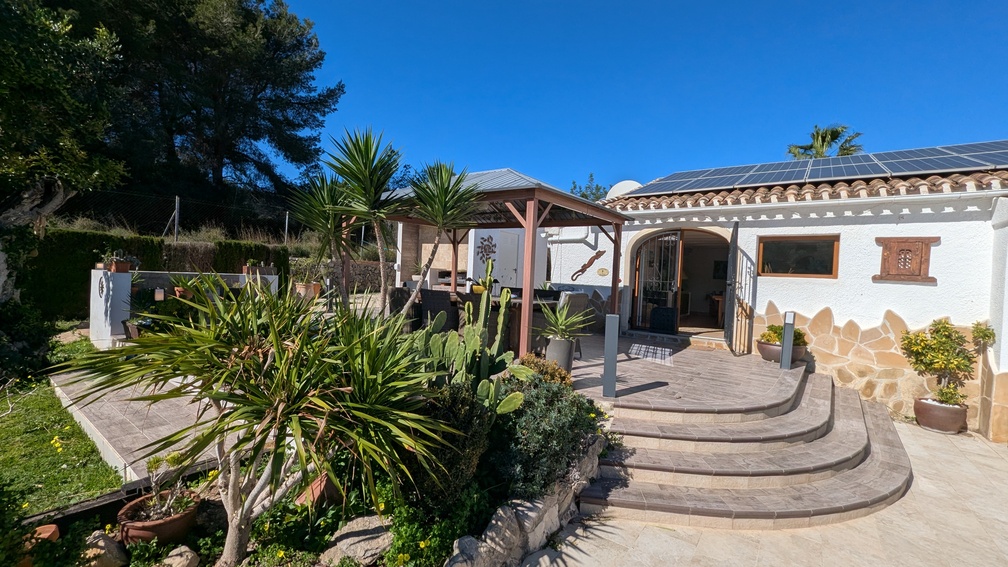 Terraza con escalones curvos de piedra, cenador de madera para comedor exterior y fachada blanca con paneles solares y vegetación mediterránea.