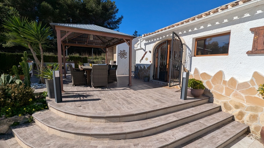 Terraza elevada con baldosas de efecto piedra y escalones curvos que dan paso a un comedor exterior bajo una pérgola de madera, junto a una fachada blanca con zócalo de piedra natural.