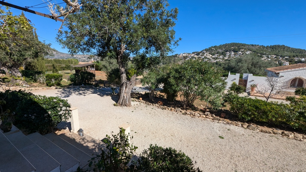 Vista elevada de la entrada de grava y los jardines mediterráneos, con olivos maduros y vistas despejadas hacia las colinas y zonas residenciales de Jávea.