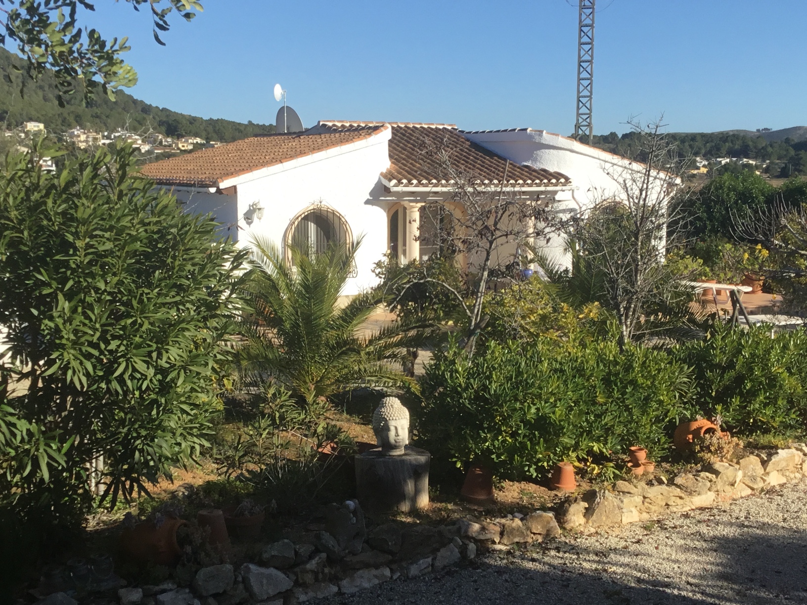Vista exterior de la villa mediterránea con fachada blanca, ventanas de arco, tejado de terracota y jardín paisajístico con palmeras y una estatua de Buda.
