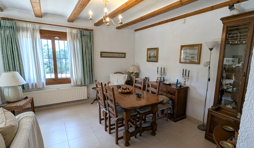 Comedor de estilo tradicional con vigas de madera a la vista, mesa de madera maciza y aparador clásico. La estancia incluye radiador y ventana con rejas de seguridad.