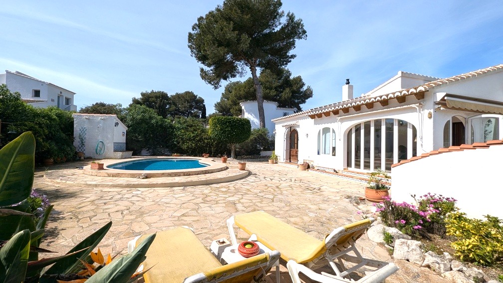 Exterior de la villa mediterránea con piscina, terraza de piedra natural y fachada blanca con vigas de madera vistas y grandes ventanales arqueados.