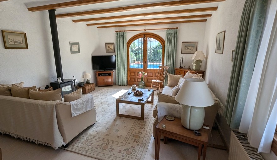 Salón con vigas de madera vistas, estufa de leña y puertas de madera en arco con acceso directo a la piscina. Mobiliario tradicional y tonos neutros crean un ambiente acogedor.