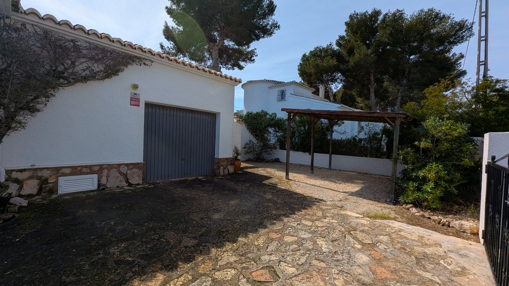 Zona de garaje y pérgola de madera con entrada de piedra natural y fachada blanca con zócalo de piedra rústica.
