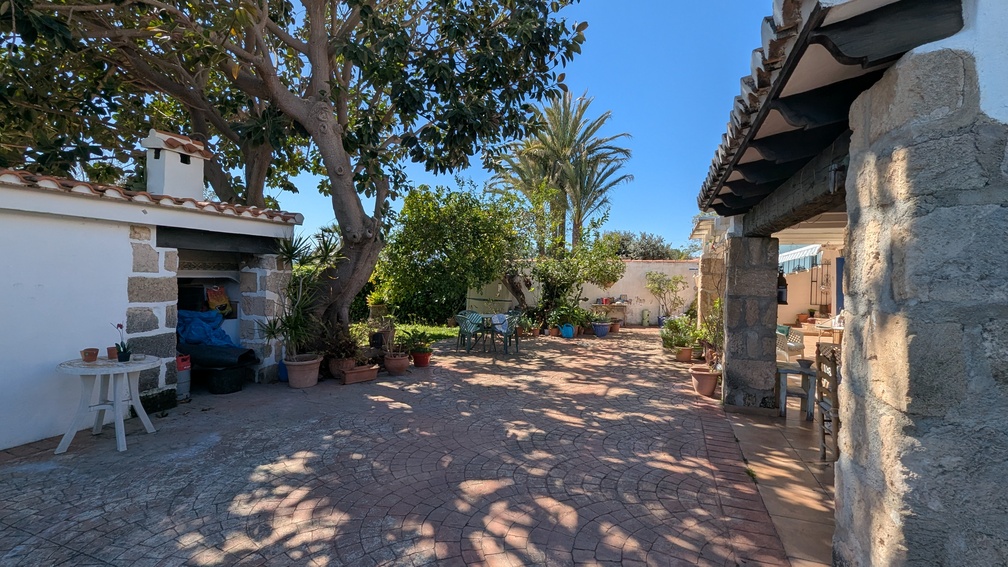 Amplio patio pavimentado con pilares de piedra Tosca, una naya sombreada y una cocina de verano exterior rodeada de árboles maduros y vegetación mediterránea.