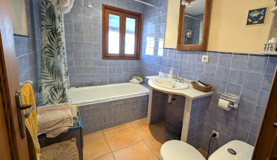 Baño tradicional con azulejos azules, bañera integrada y lavabo con encimera de mármol. Incluye suelos de terracota y una ventana de madera que aporta luz natural.