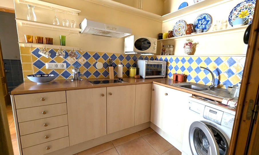 Cocina compacta en forma de L con salpicadero de azulejos en rombos azules y amarillos, encimeras de madera, estanterías abiertas y electrodomésticos integrados.