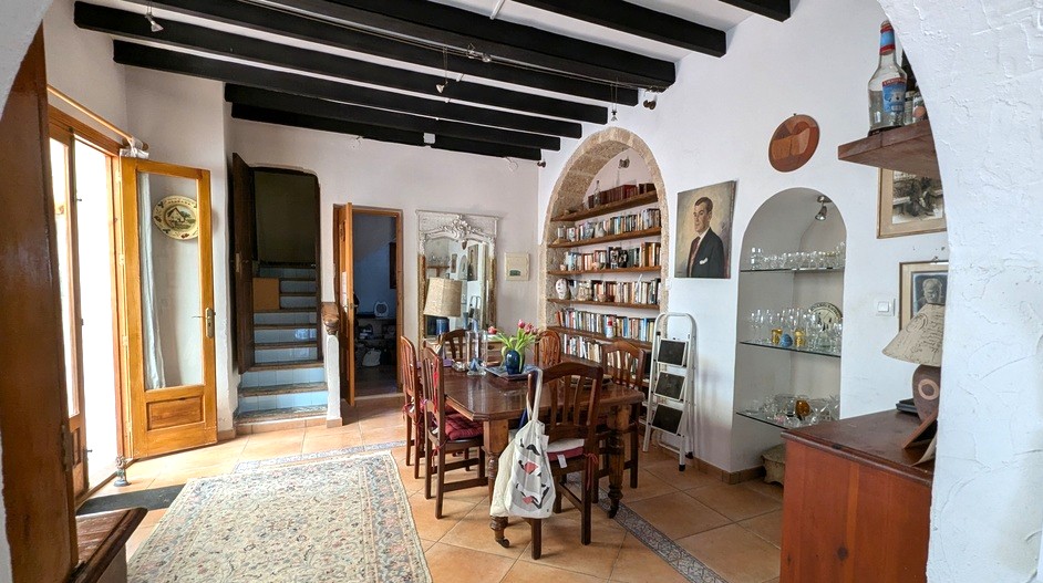 Comedor tradicional con vigas de madera oscura, estantería integrada en arco de piedra Tosca y suelos de barro cocido junto a una escalera decorada con azulejos.