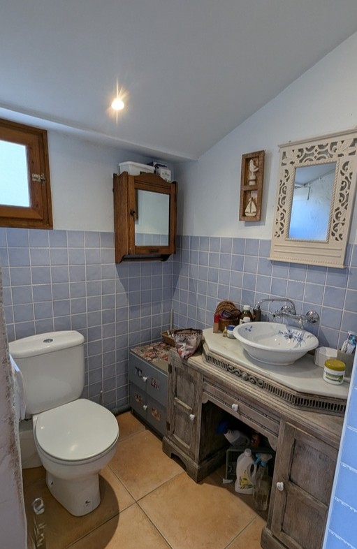 Cuarto de baño con lavabo cerámico de motivos florales sobre mueble de madera rústica, encimera de mármol, azulejos en azul suave y suelo de barro.