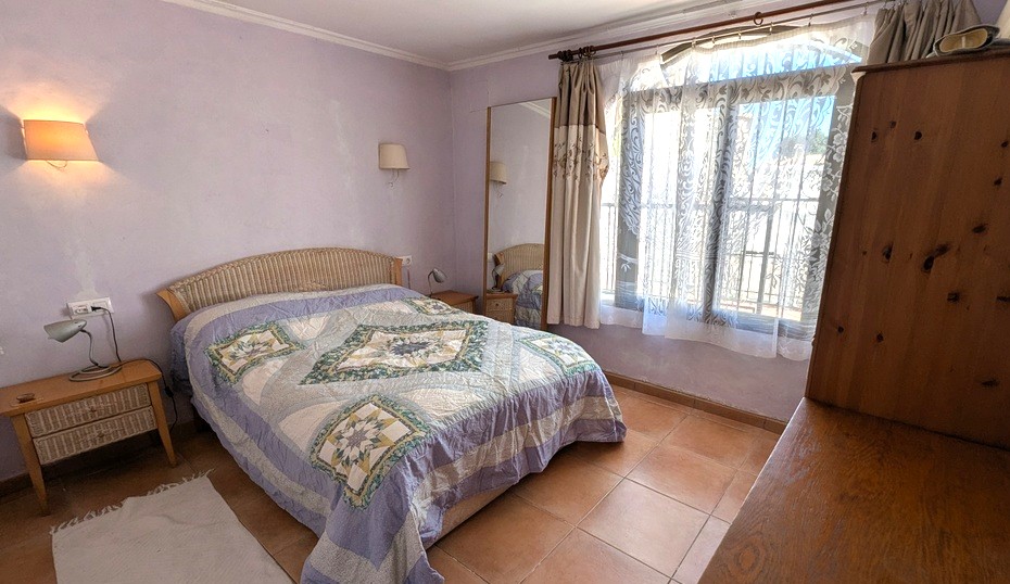 Dormitorio con paredes en tono lavanda, suelos de gres rústico, cama con cabecero de mimbre y un gran ventanal que aporta abundante luz natural.