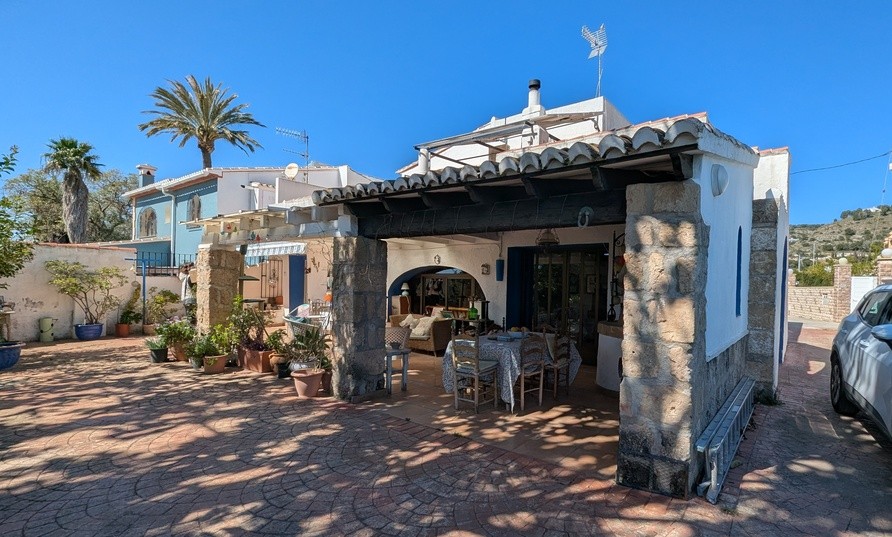Naya tradicional con pilares de piedra Tosca y vigas de madera, que incluye zona de comedor exterior y estar frente al patio pavimentado con plantas.