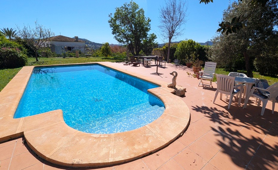 Piscina privada con revestimiento de gresite azul, rodeada de una amplia terraza de terracota y un jardín mediterráneo con vistas despejadas a las montañas.