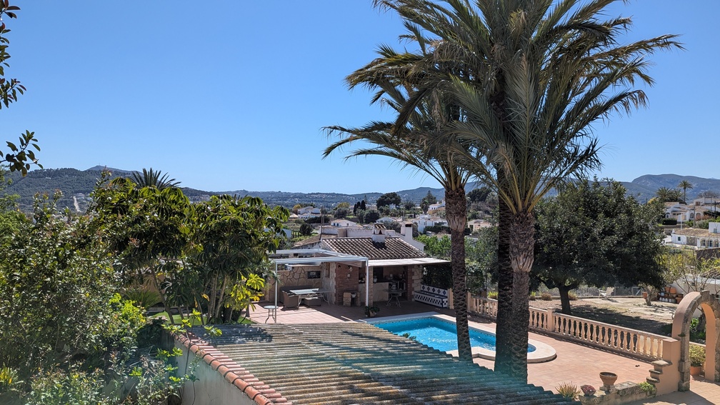 Vista elevada de la piscina y terraza, con una cocina de verano tradicional, palmeras maduras y vistas despejadas al paisaje rural de Jávea.