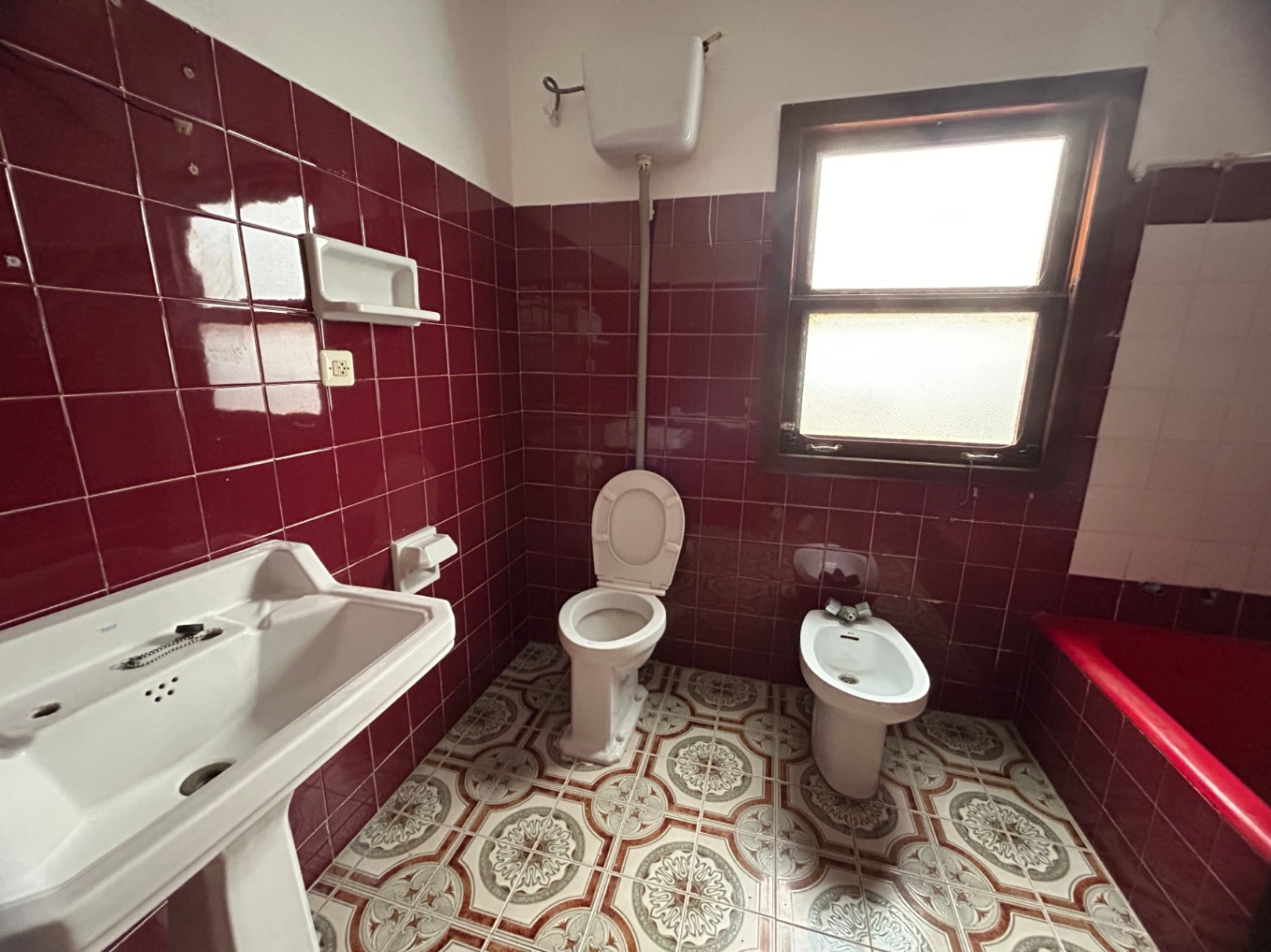 Baño original con azulejos granate, suelo hidráulico decorado y sanitarios clásicos. Dispone de ventana de madera y luz natural, ideal para un proyecto de reforma integral.