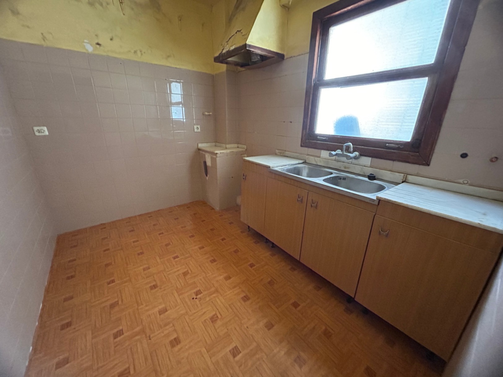 Cocina original con fregadero de doble seno, mobiliario de madera y suelos cerámicos. Dispone de ventana exterior y campana antigua, ideal para un proyecto de reforma integral.