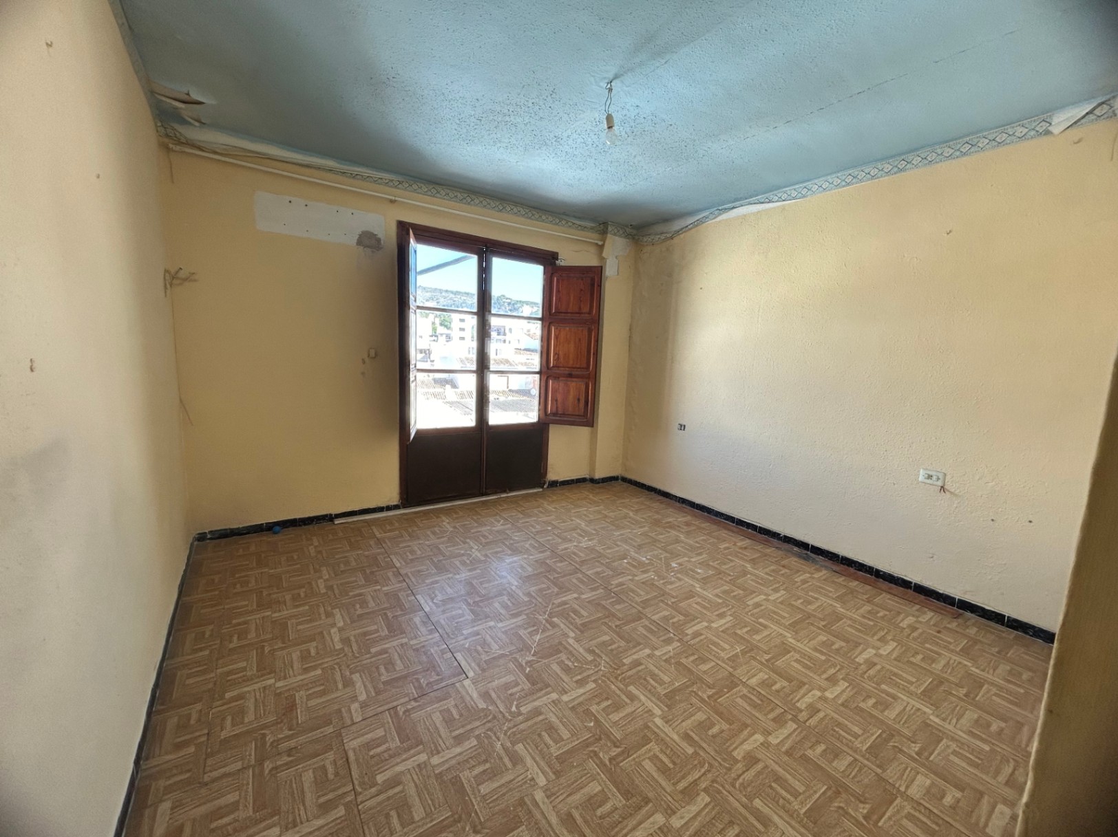 Habitación para reformar con suelo de gres cerámico estampado, paredes en tono crema y acceso directo a un balcón mediante puertas de madera con contraventanas.