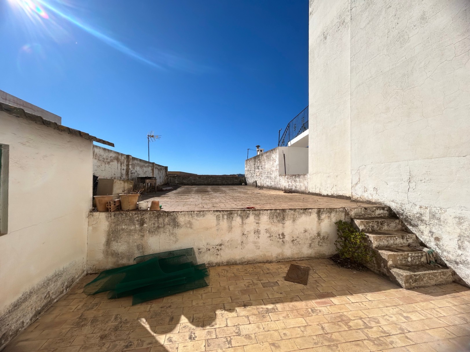 Terraza comunitaria en azotea con dos niveles pavimentados, conectada por escaleras de piedra y rodeada de muros blancos bajo un cielo despejado.