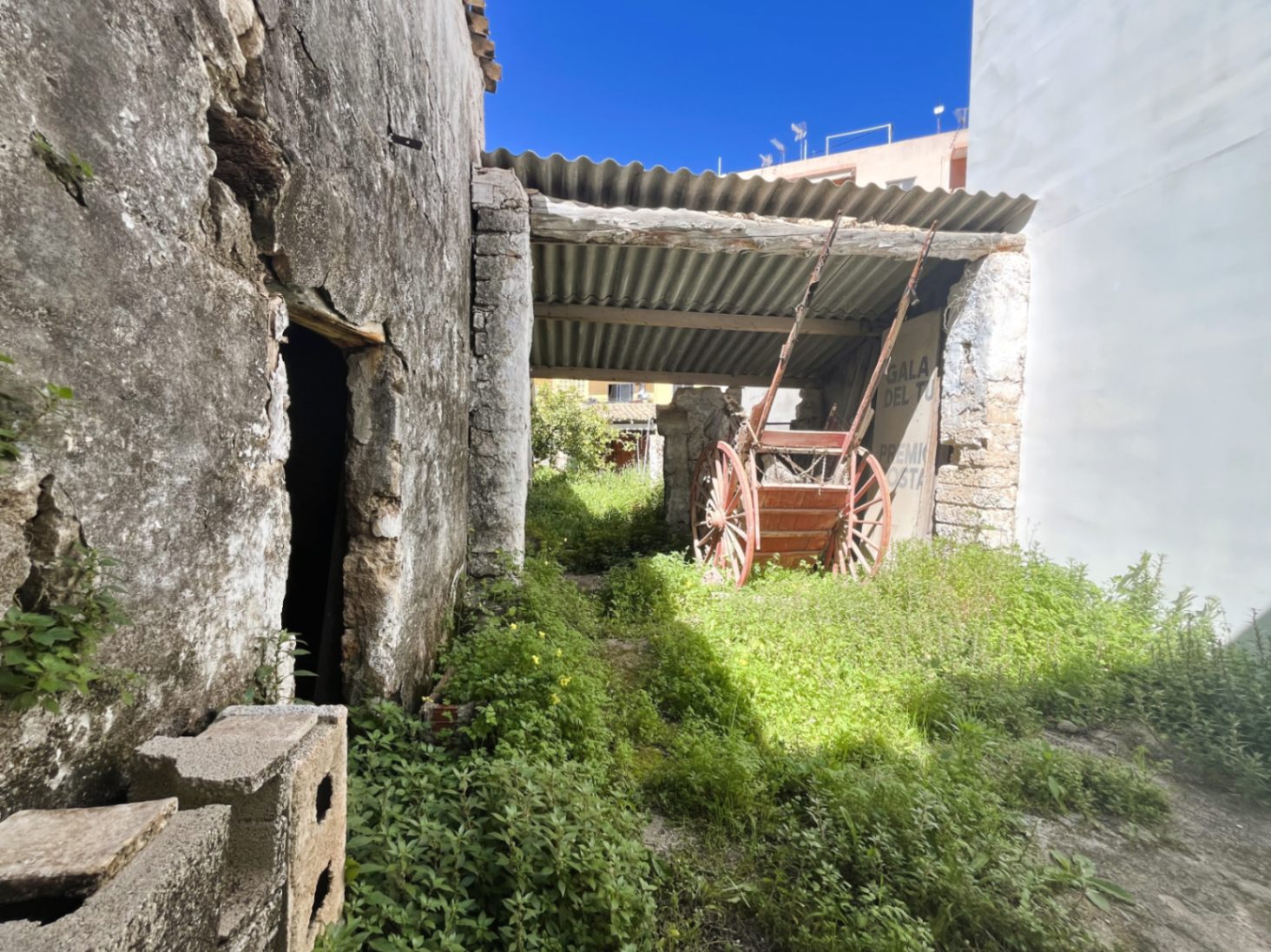 Patio trasero rústico con muros de piedra original, vegetación natural y un carro antiguo de madera bajo una estructura techada, reflejando el carácter histórico de la propiedad.