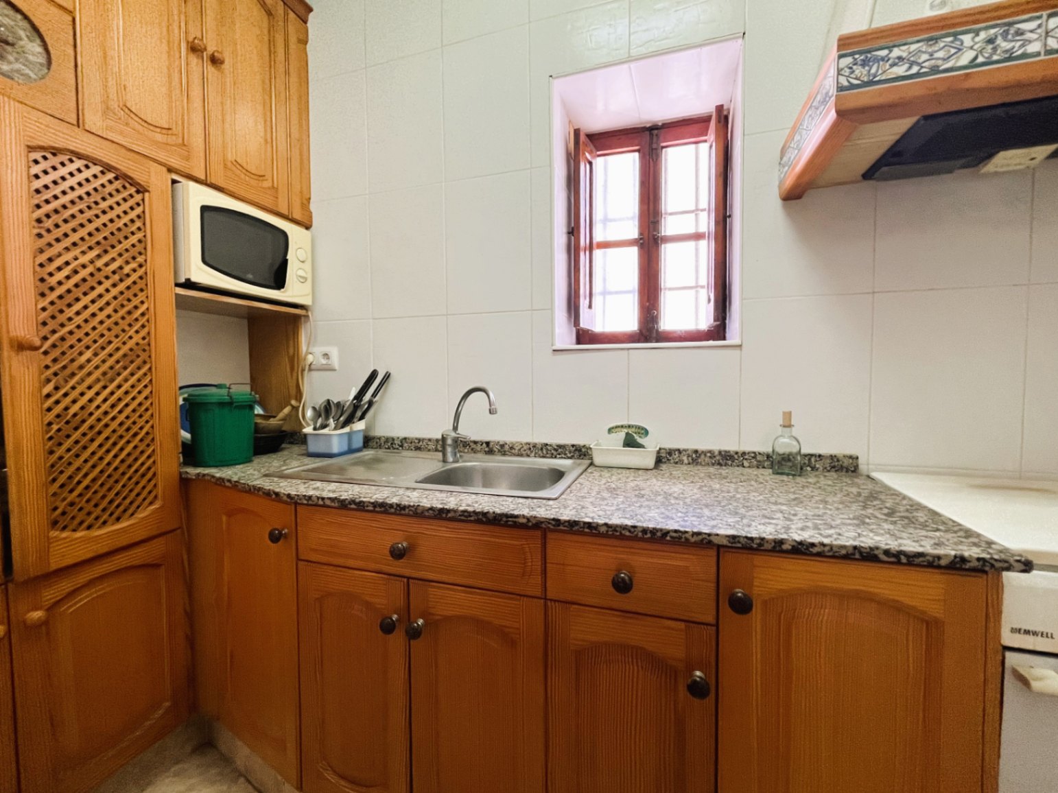Cocina cerrada con armarios de madera tradicional, encimera de granito y fregadero doble, iluminada por una ventana con contraventanas de madera originales.