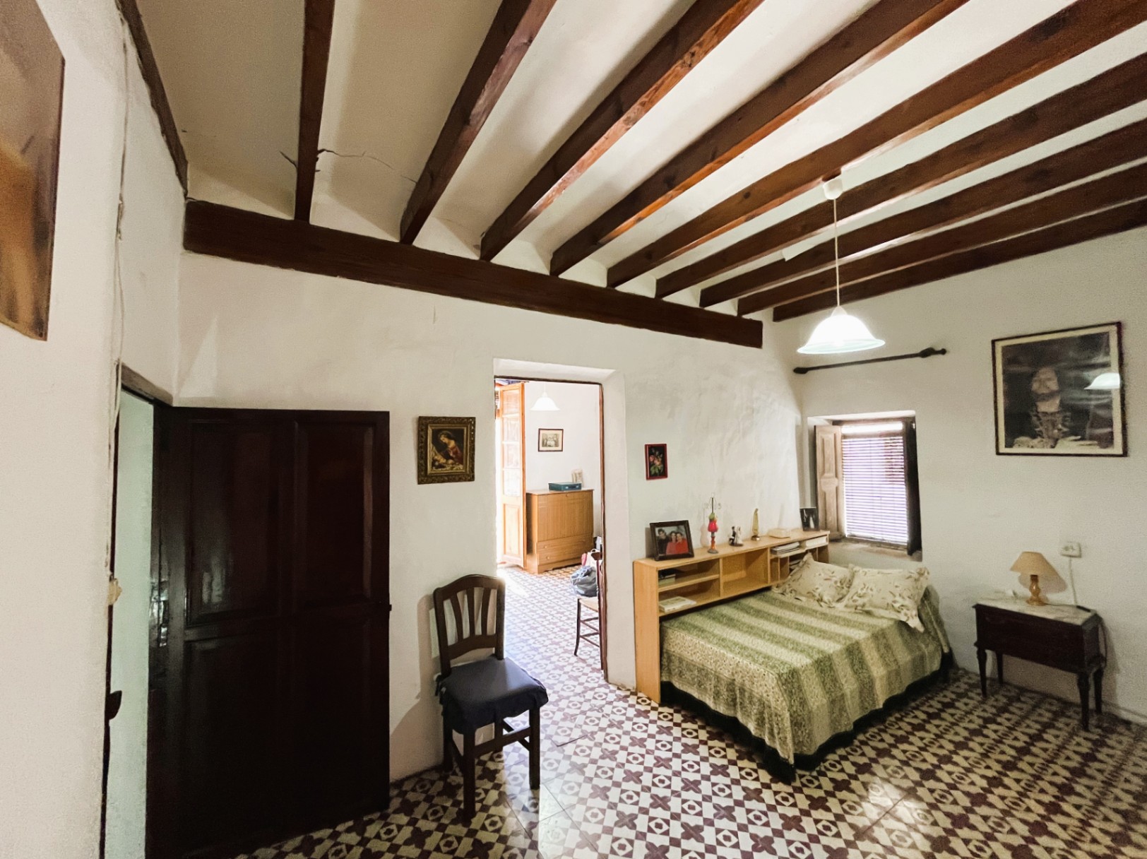 Dormitorio con vigas de madera vista, suelos de baldosa hidráulica original y muros de yeso blanco. Presenta una ventana con contraventanas y acceso directo a otras estancias.