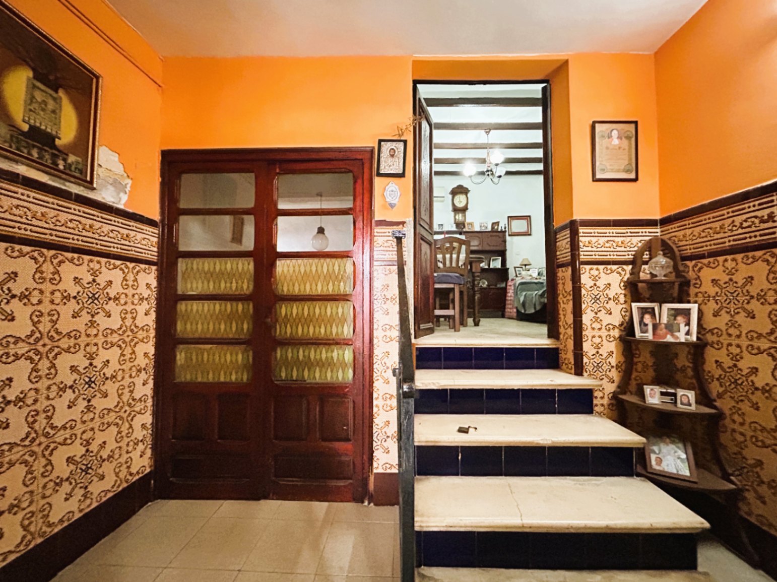 Entrada tradicional con zócalos de azulejos ornamentados, paredes en tono terracota y escalera de mármol con contrahuellas azules que conduce a estancias con vigas de madera.