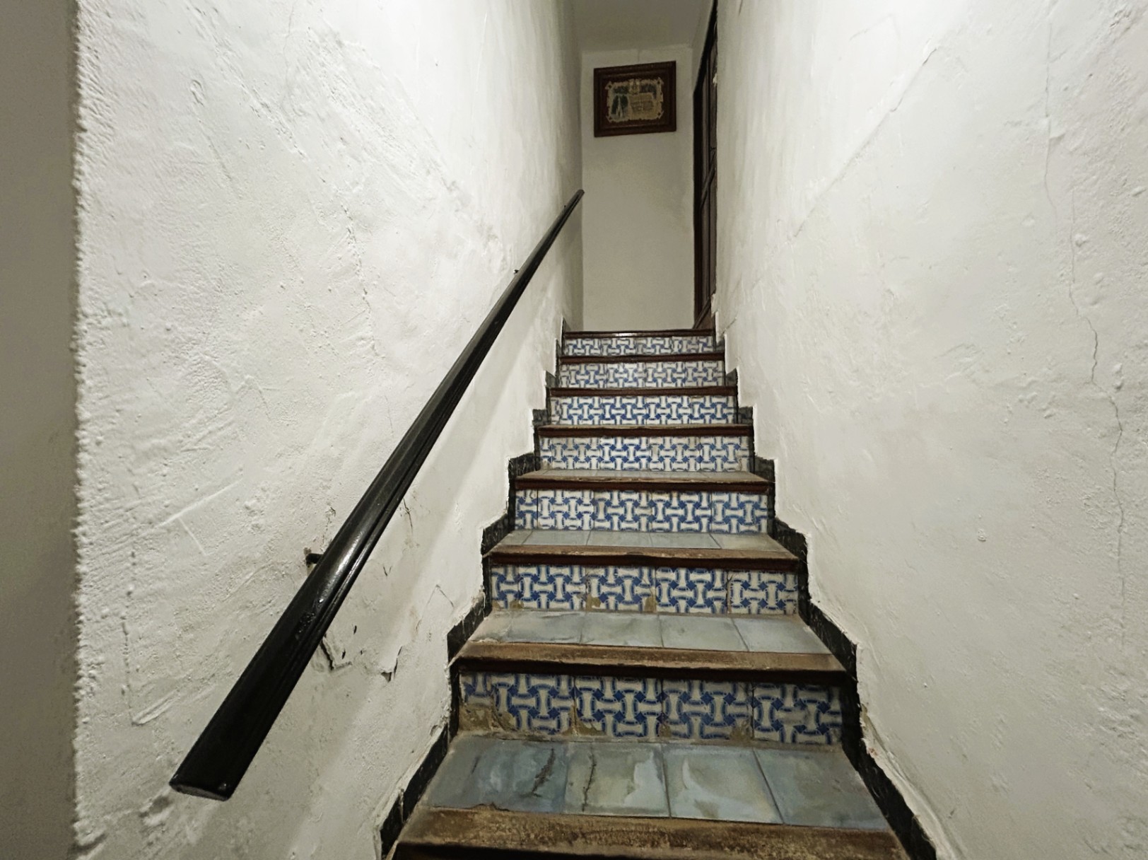 Escalera tradicional con contrahuellas decoradas con azulejos en azul y blanco, peldaños de madera y pasamanos oscuro sobre paredes blancas de estilo rústico.