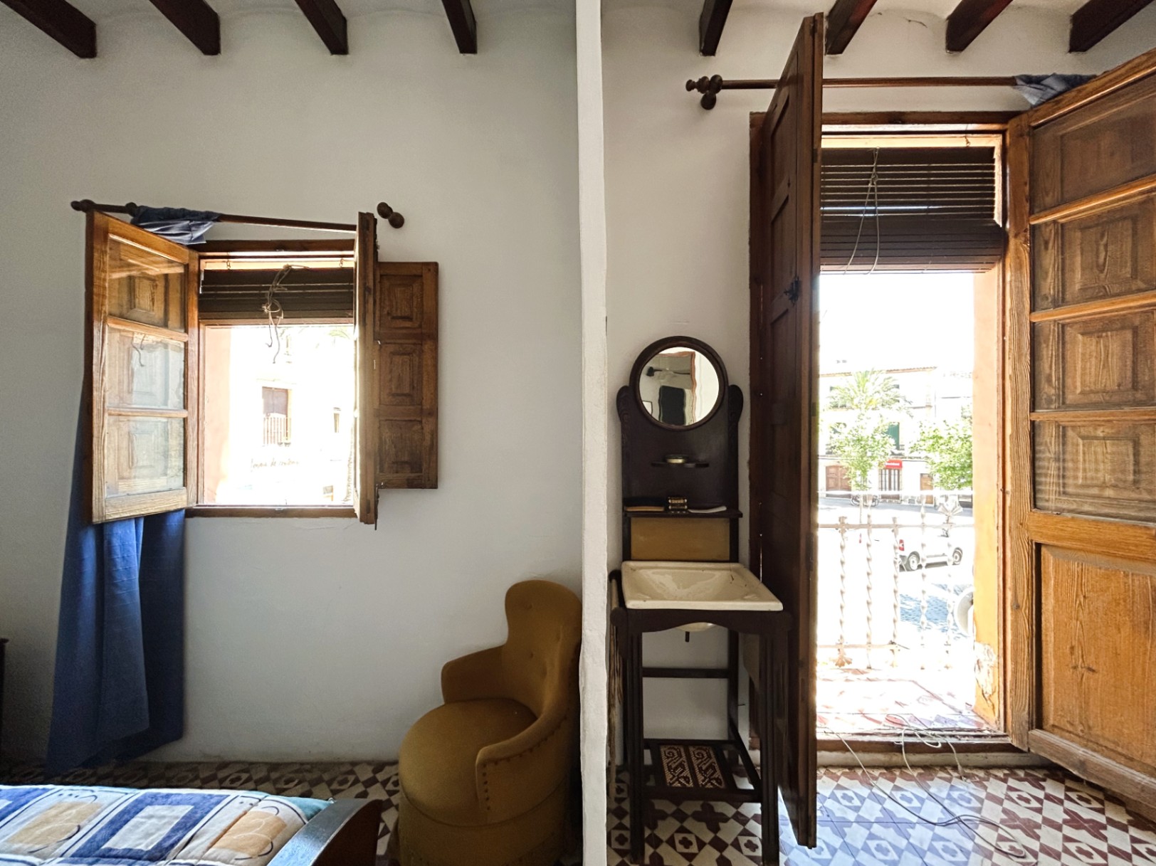 Habitación tradicional con vigas de madera, suelos hidráulicos originales y acceso a un balcón con vistas al casco antiguo de Jávea. Incluye un lavabo de época.