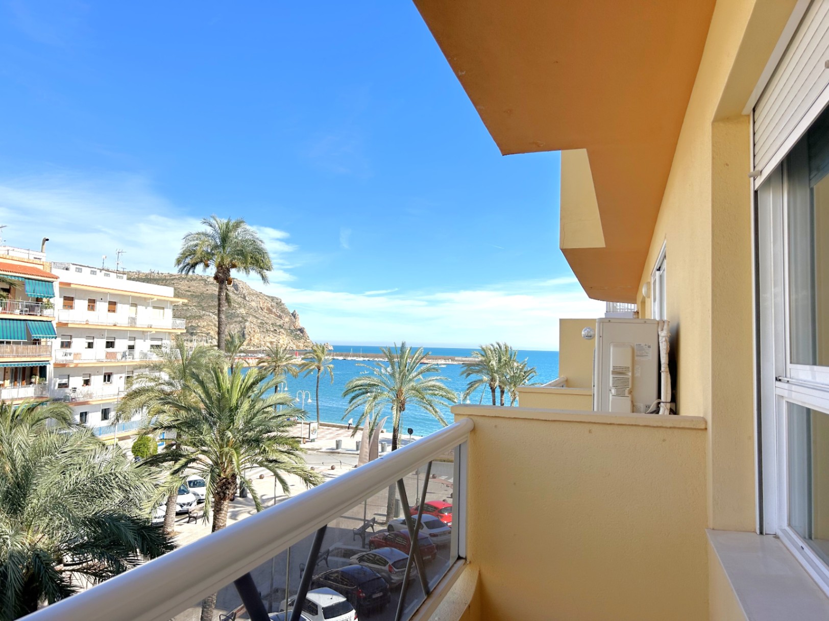 Balcón con vistas directas al mar y al Puerto de Jávea, destacando el entorno costero con palmeras y carpintería exterior de aluminio blanco.