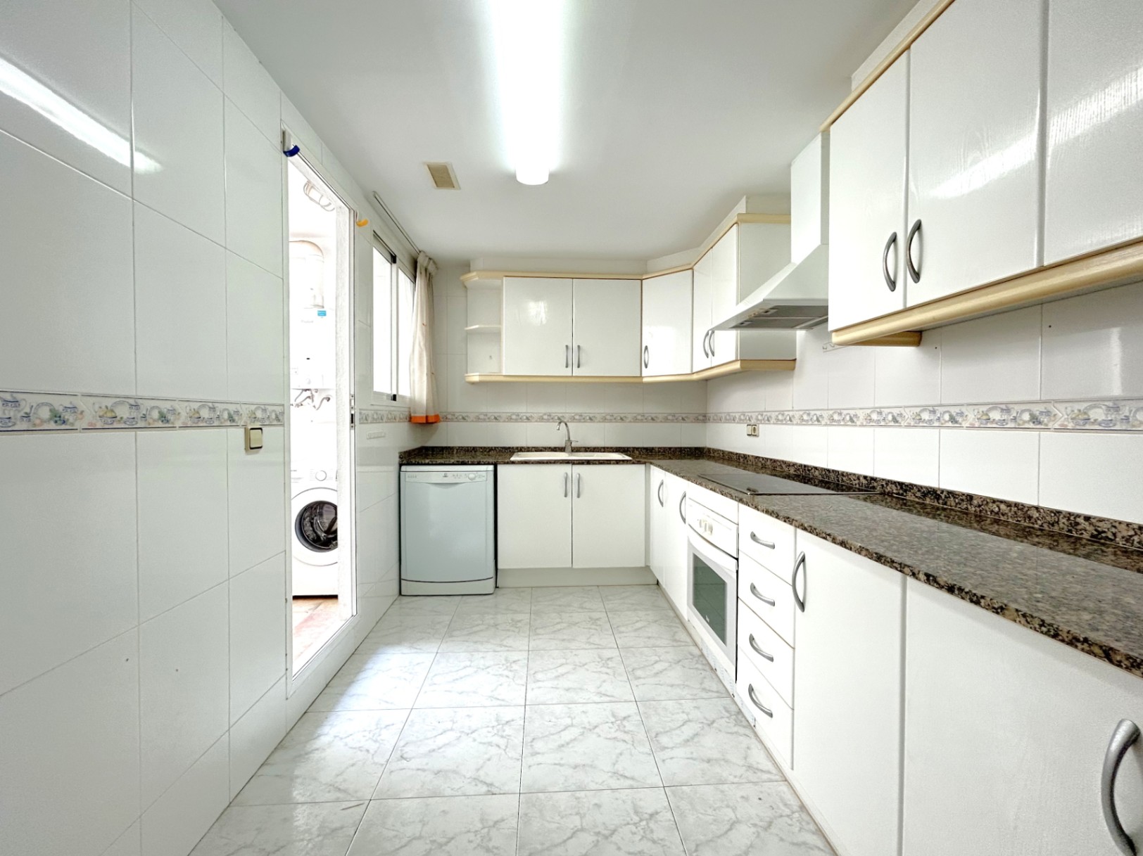 Cocina independiente en forma de L con armarios blancos, encimera de granito oscuro y acceso directo a una zona de lavadero exterior.