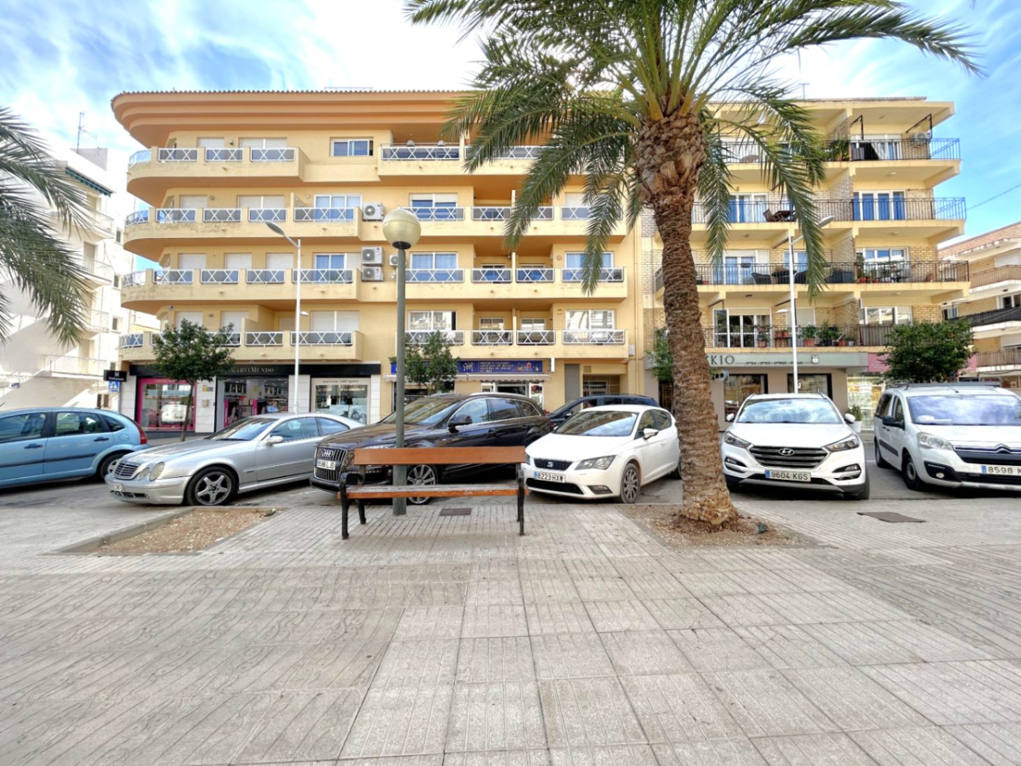 Vista exterior del edificio en el Puerto de Jávea, con fachada ocre y balcones con barandillas blancas frente a una plaza peatonal con palmeras.