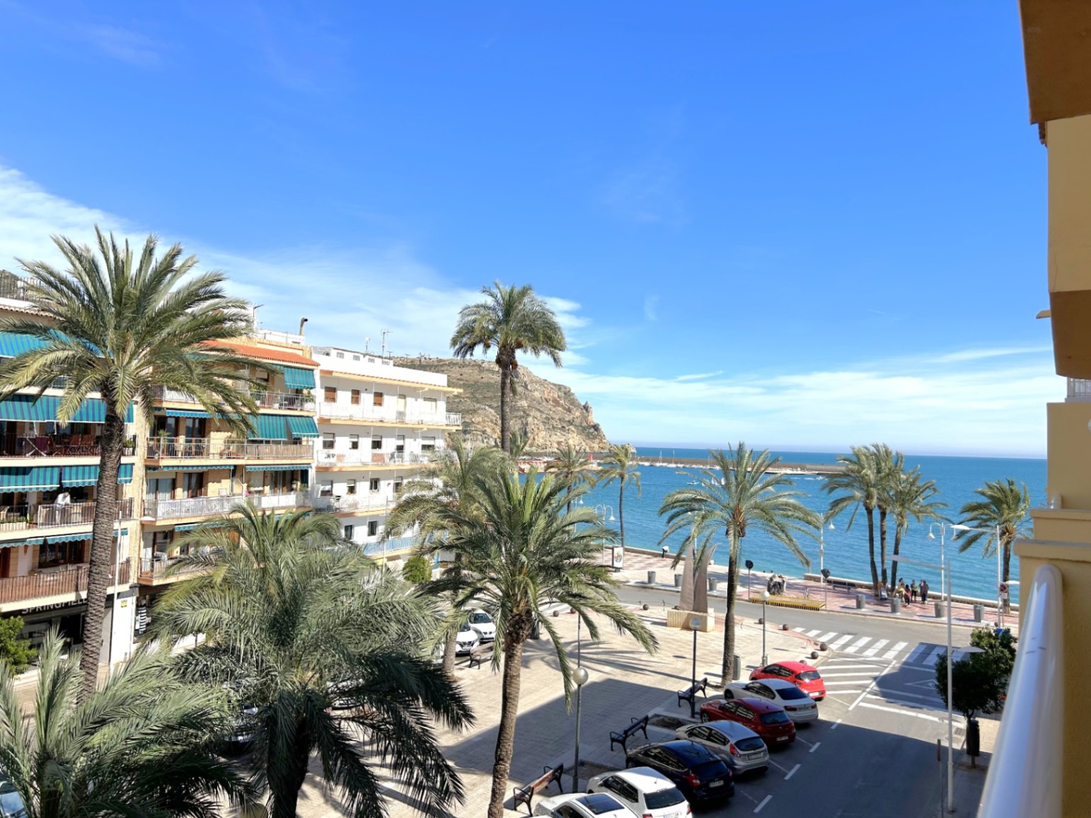 Vistas desde el balcón al paseo marítimo del Puerto de Jávea, con palmeras, el mar Mediterráneo y el cabo al fondo bajo un cielo despejado.