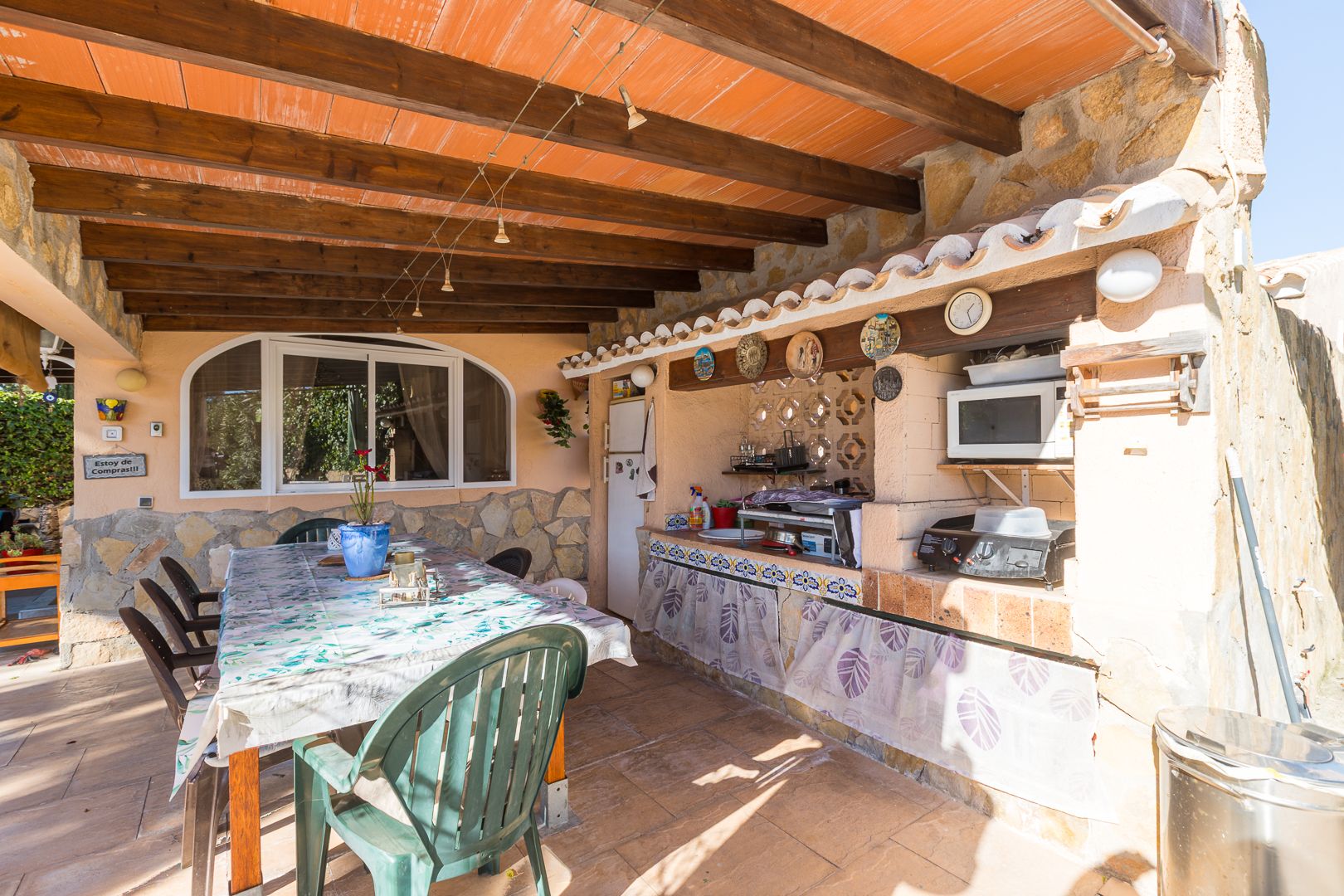 Cocina de verano y comedor exterior bajo un porche con vigas de madera vista, paredes de piedra natural y zona de barbacoa funcional.