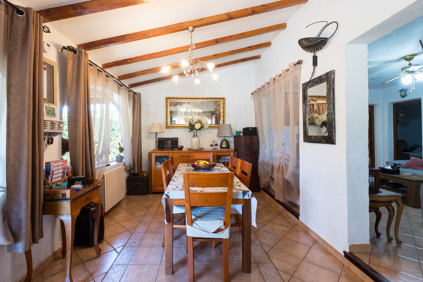 Comedor luminoso con techo inclinado de vigas de madera vistas, suelo de gres rústico y mobiliario de madera clásica, conectado visualmente con el salón.