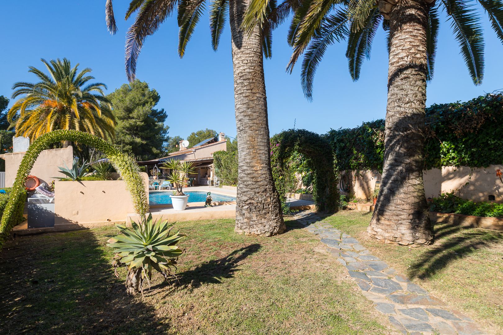 Jardín mediterráneo con grandes palmeras, camino de piedra natural y vista hacia la piscina y la villa con placas solares.