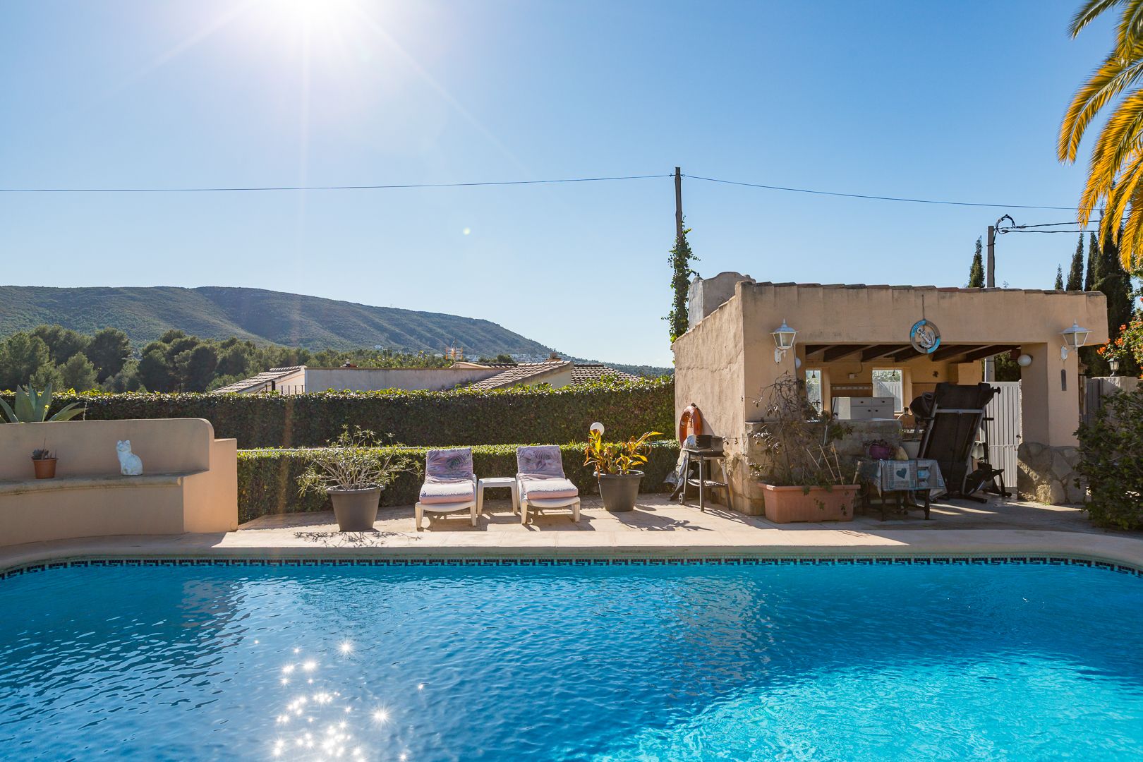 Piscina con borde de mosaico, zona de solárium con hamacas y cocina de verano cubierta, con vistas despejadas a la montaña bajo el sol mediterráneo.