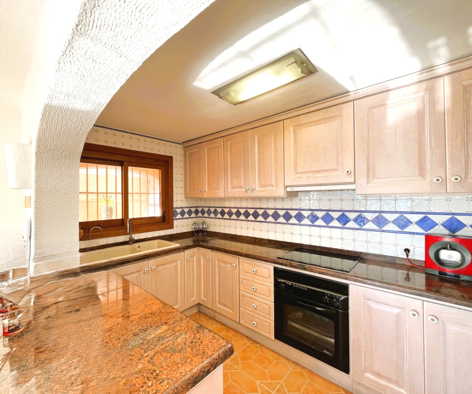 Cocina equipada con armarios de madera clara, encimera de granito y salpicadero de azulejos con detalles azules. Incluye horno integrado, placa vitrocerámica y ventana de madera.