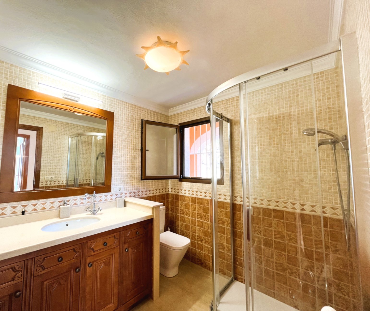 Cuarto de baño con mueble de madera tallada, encimera blanca, ducha de esquina con mampara curva y paredes revestidas en azulejos de tonos tierra.