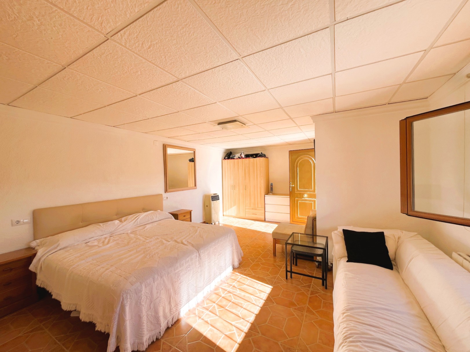 Dormitorio con suelos de terracota, techo de placas y zona de estar integrada. Incluye mobiliario de madera y recibe luz natural directa sobre el pavimento cerámico.