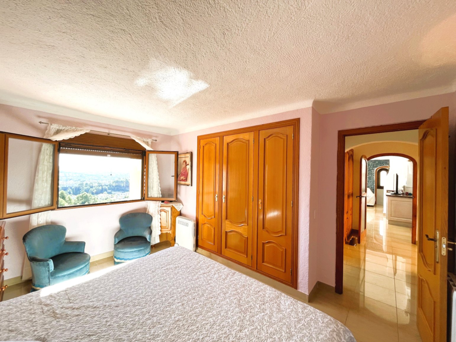 Dormitorio luminoso con armarios empotrados de madera, zona de estar con butacas y ventana con vistas panorámicas despejadas al valle.