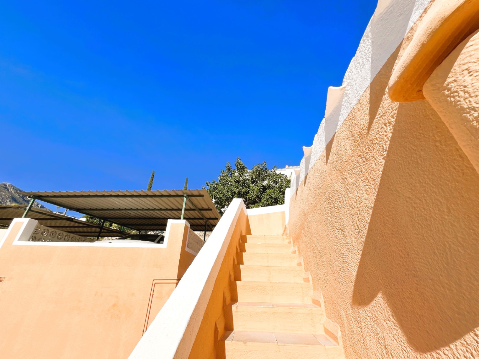 Escalera exterior de baldosas cerámicas flanqueada por muros en tonos tierra y blanco, situada junto a la zona de aparcamiento cubierto con pérgola metálica.