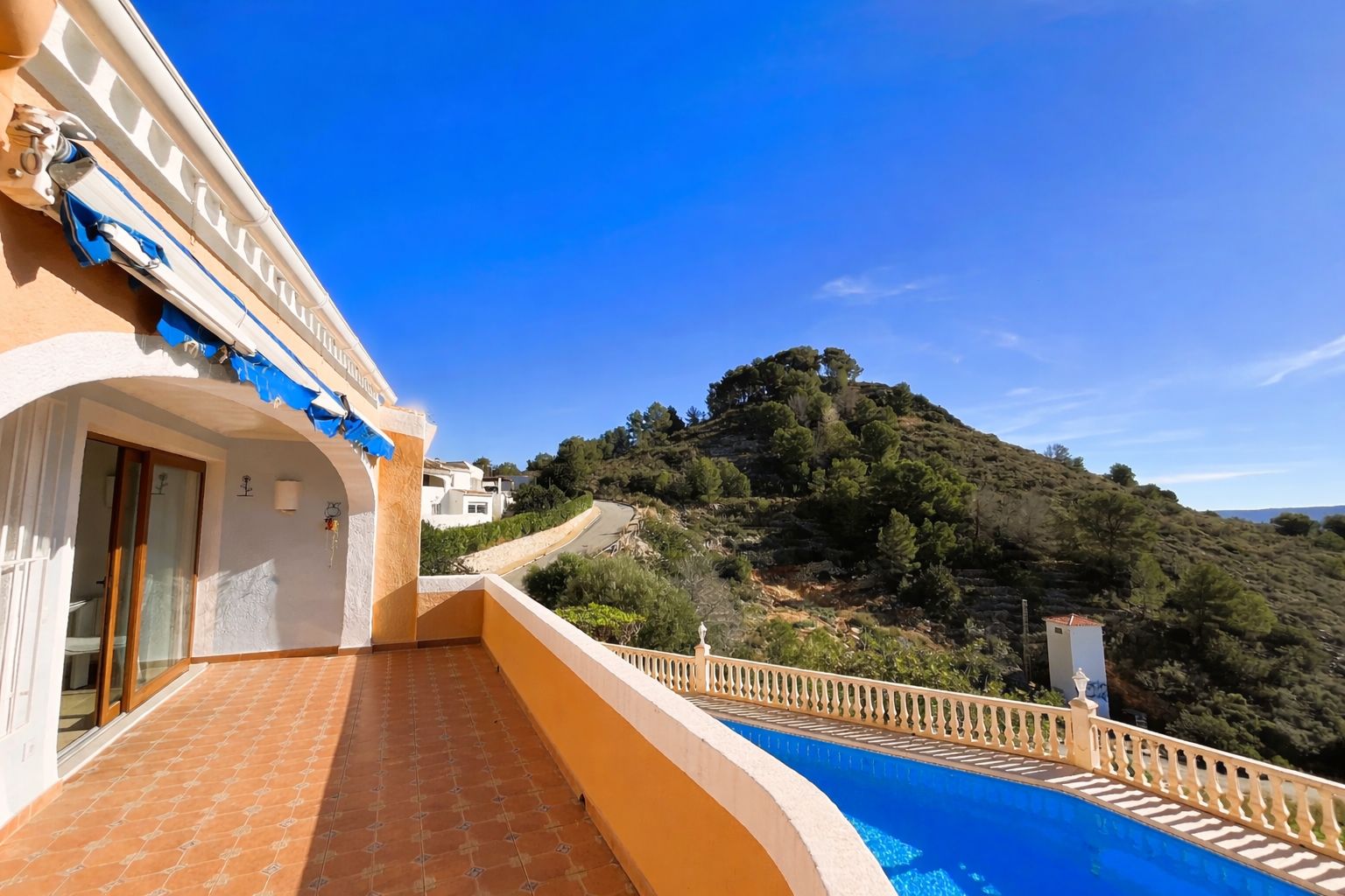 Terraza superior con suelo de gres, toldo extensible y vistas despejadas a la montaña y la piscina inferior en un entorno natural.