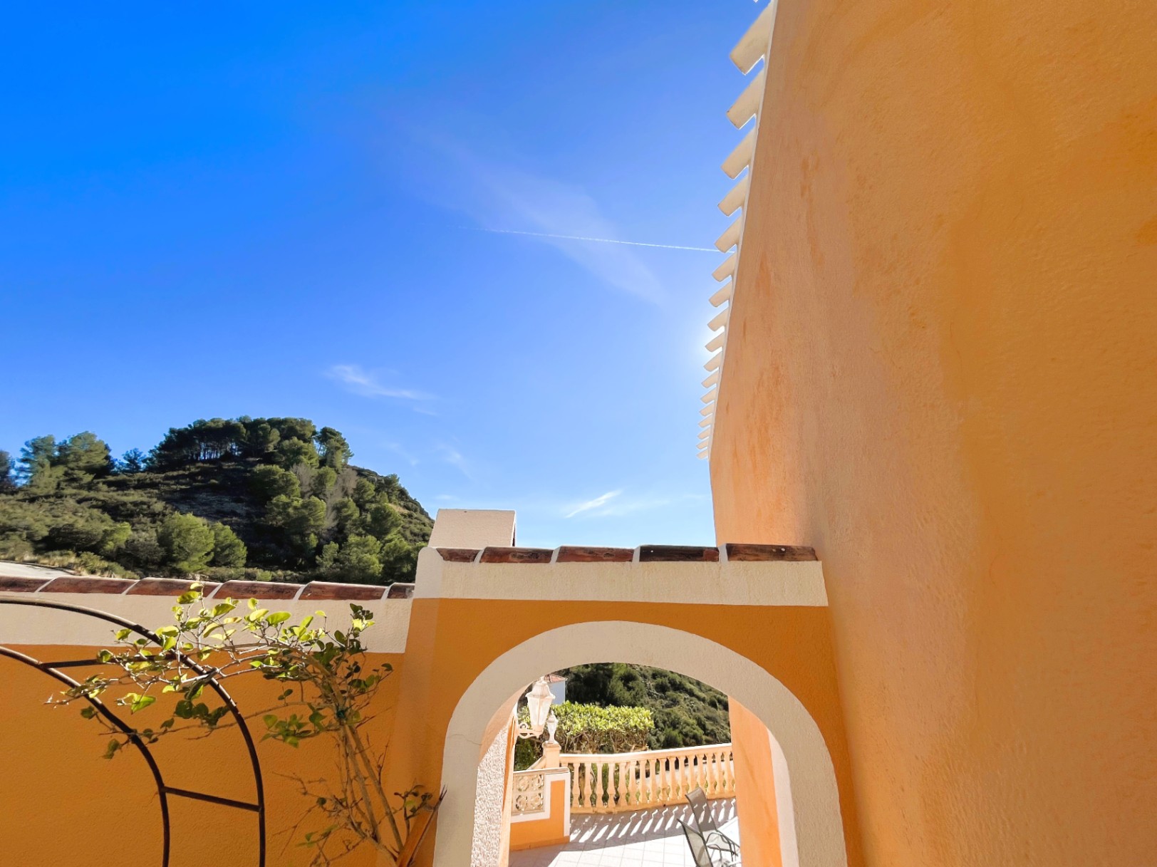 Vista exterior que muestra un arco de entrada blanco sobre fachada ocre, con detalles en teja cerámica y vistas a las colinas arboladas del Montgó.