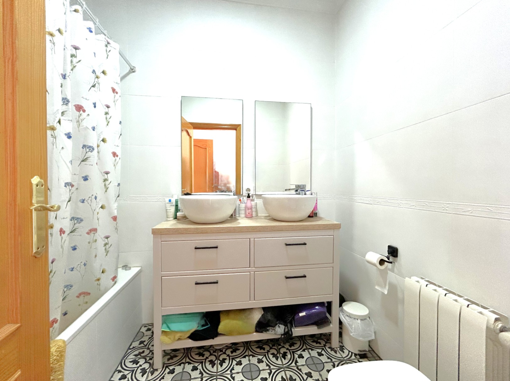 Baño completo con mueble de doble lavabo sobre encimera de madera, suelos de baldosa hidráulica decorativa, bañera y radiador de calefacción central.