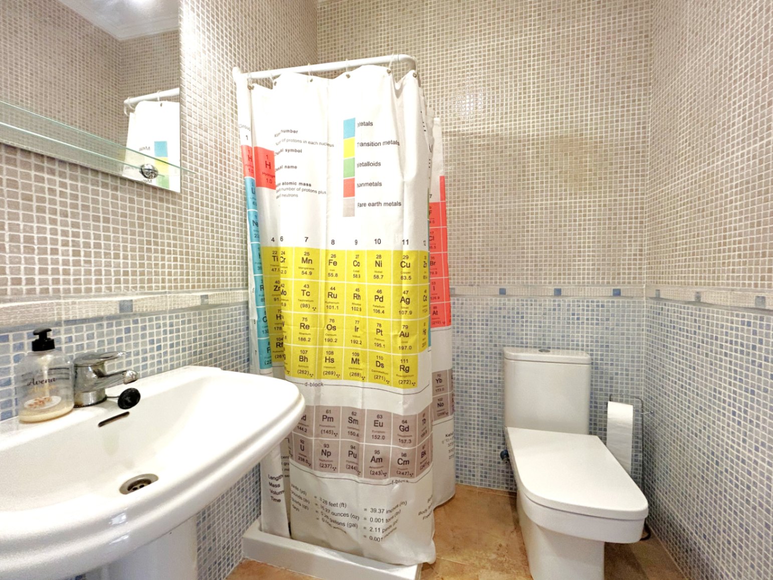Baño con paredes revestidas en mosaico bitono, lavabo de porcelana blanca y una original cortina de ducha con la tabla periódica. Incluye inodoro compacto y espejo con repisa de cristal.
