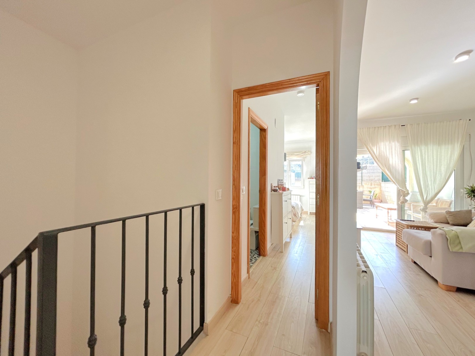 Distribuidor luminoso con suelos de madera y barandilla de hierro forjado que conecta el salón, los dormitorios y el baño, mostrando un radiador de calefacción central.