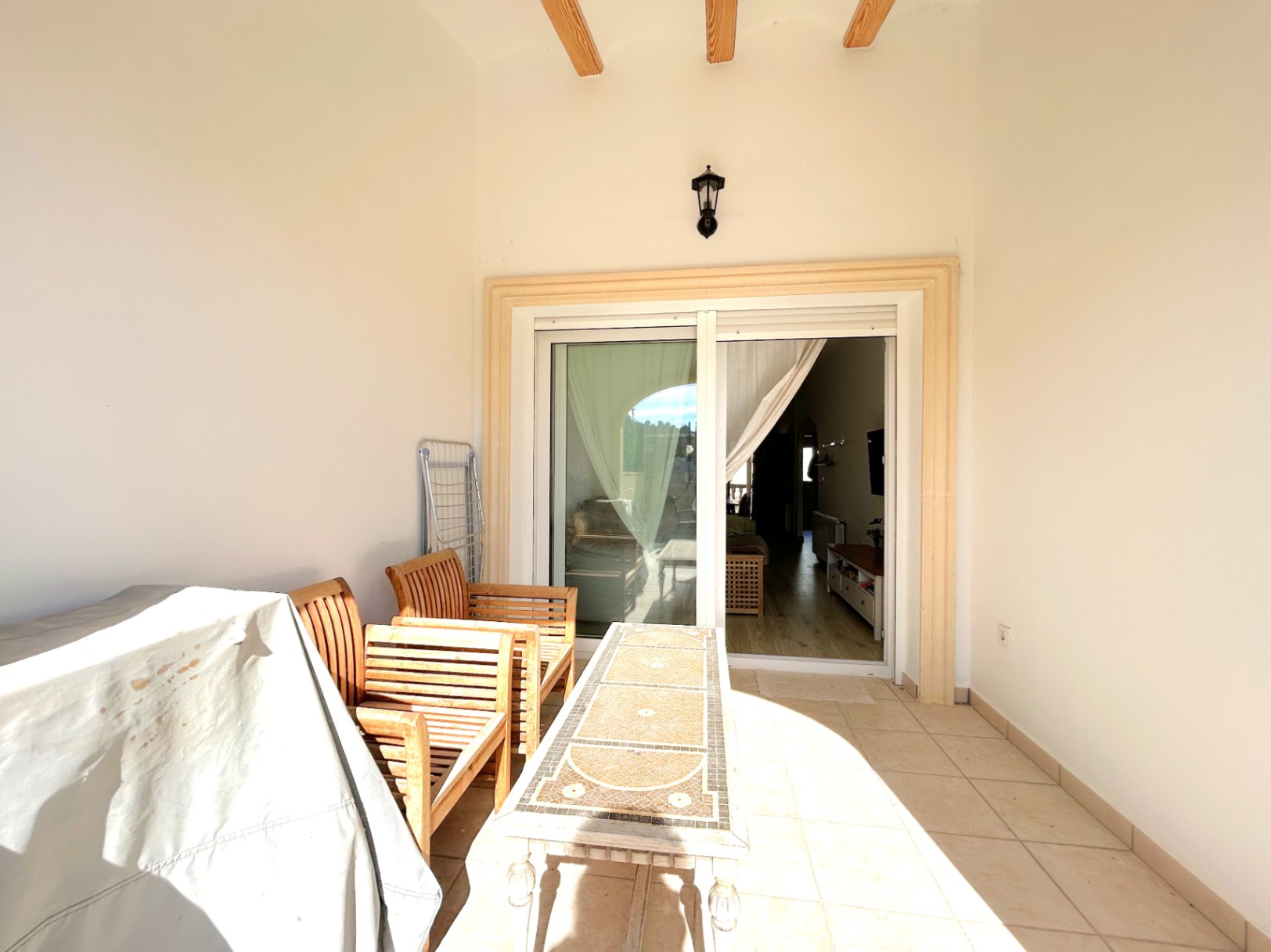 Terraza cubierta con vigas de madera a la vista, suelo cerámico y mobiliario. El ventanal corredero conecta directamente el exterior con el salón comedor.