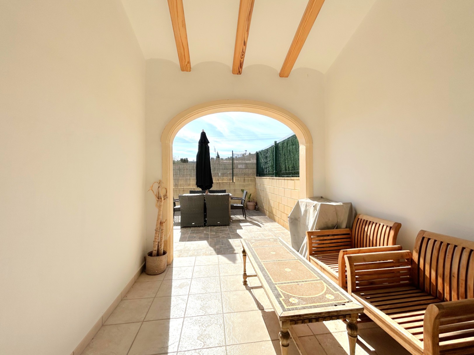 Terraza cubierta con vigas de madera a la vista y arco de piedra, equipada con mobiliario de madera y acceso directo a la zona exterior soleada.