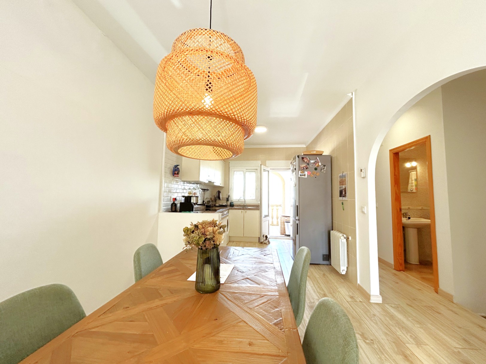 Zona de comedor con mesa de madera geométrica y lámpara de mimbre, integrada con cocina abierta blanca y acceso directo al lavadero exterior.